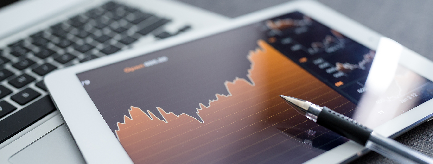 Close-up of a digital tablet displaying a financial graph or stock market chart with a pen resting on the screen, positioned next to a laptop keyboard