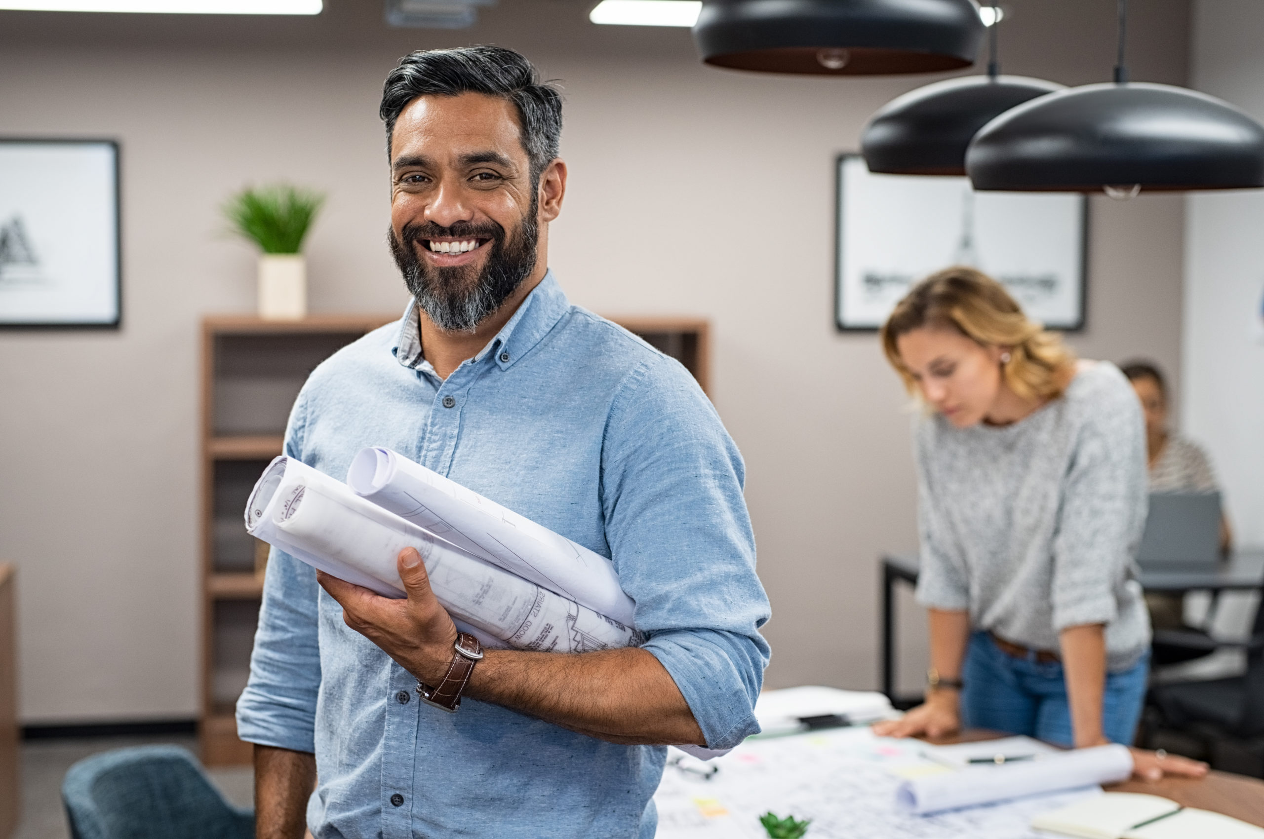 A waist-up portrait of a smiling man with a salt-and-pepper beard and dark hair standing in a modern office