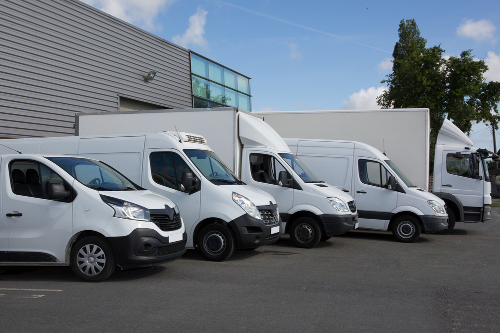 A diagonal row of white commercial delivery vehicles, ranging from smaller panel vans in the foreground to larger box trucks in the background, parked on an asphalt lot in front of a grey industrial warehouse building under a blue sky with scattered clouds