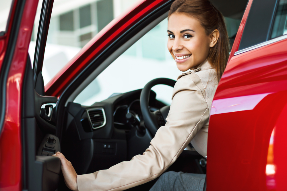 A smiling woman sitting in the driver's seat of a red car with the door open, looking back over her shoulder at the camera