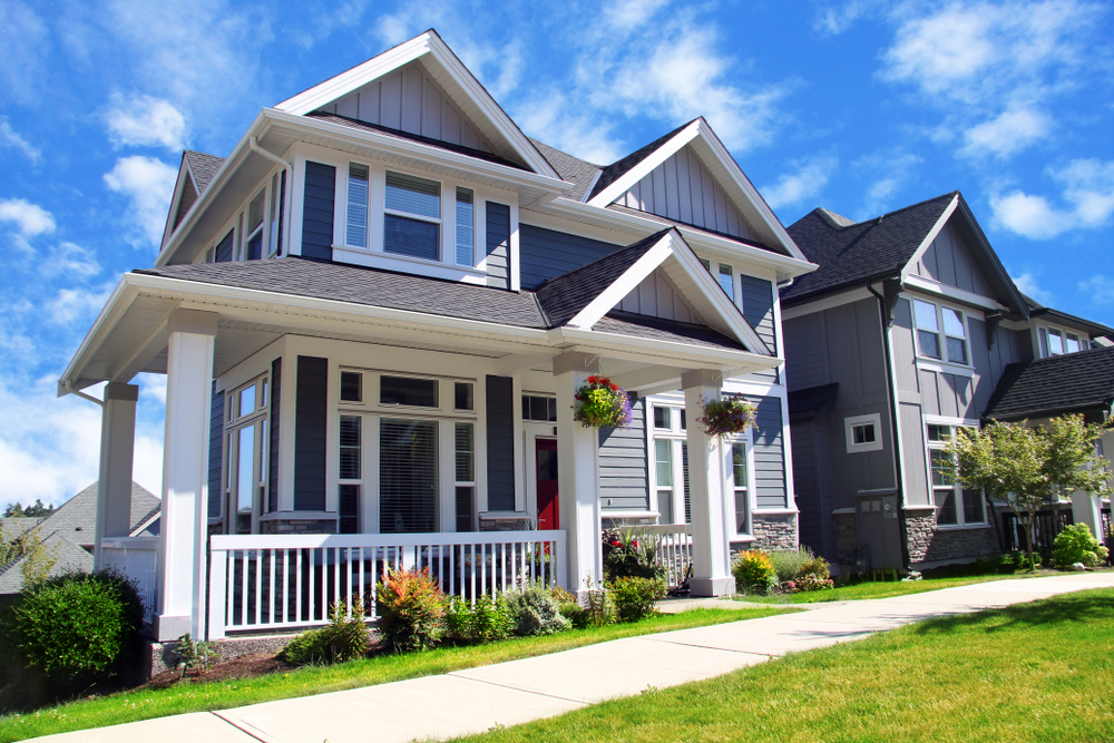 A large, two-story suburban home features blue-gray siding, white trim, and multiple gabled roof peaks under a bright blue sky with scattered clouds