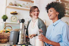 Two women smile together in a kitchen, standing in front of a blender filled with ingredients Two women smile together in a kitchen, standing in front of a blender filled with ingredients