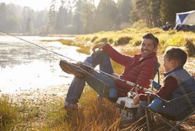 Two men sitting in chairs, fishing by a serene lake surrounded by trees and calm waters Two men sitting in chairs, fishing by a serene lake surrounded by trees and calm waters